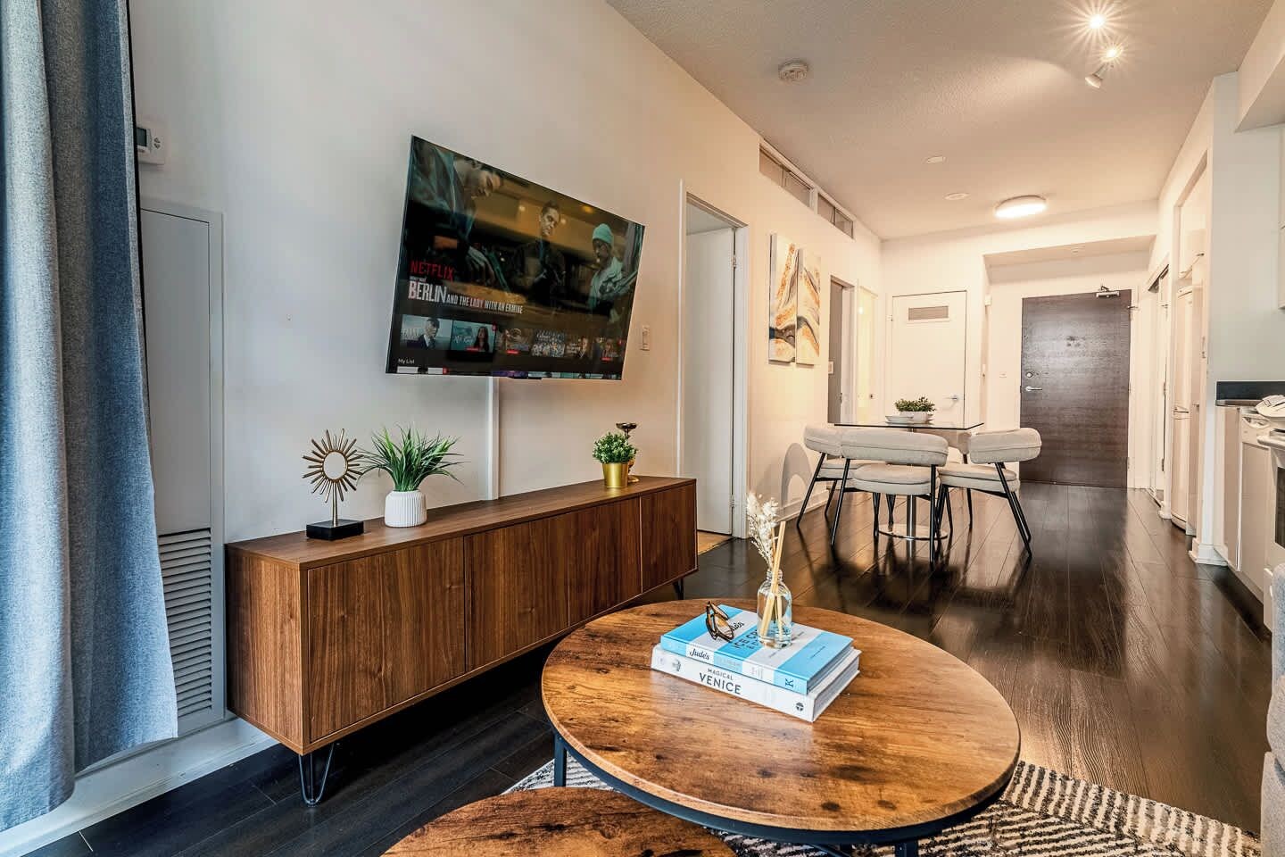 A modern living room with a round wooden coffee table, a mounted TV, and a dining area with a white table and chairs.