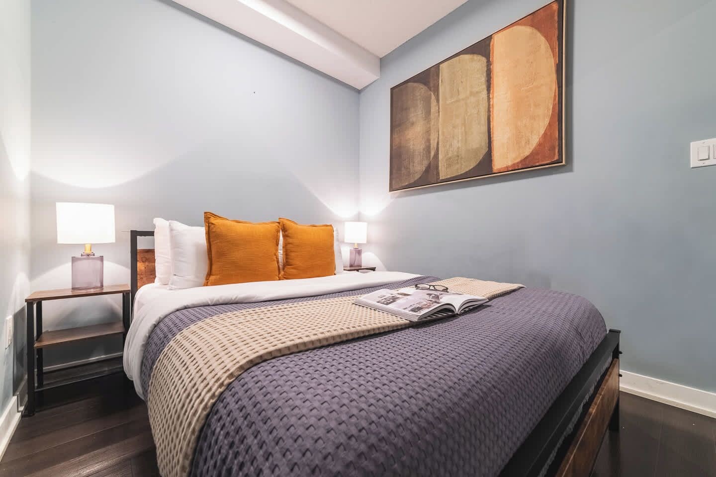 A bedroom with a bed, two bedside tables with lamps, a large abstract painting on the wall, and a magazine on the bed.