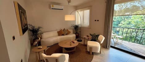 A modern living room with a sofa, coffee table, and chairs, a ceiling fan, and a balcony with a view of greenery.