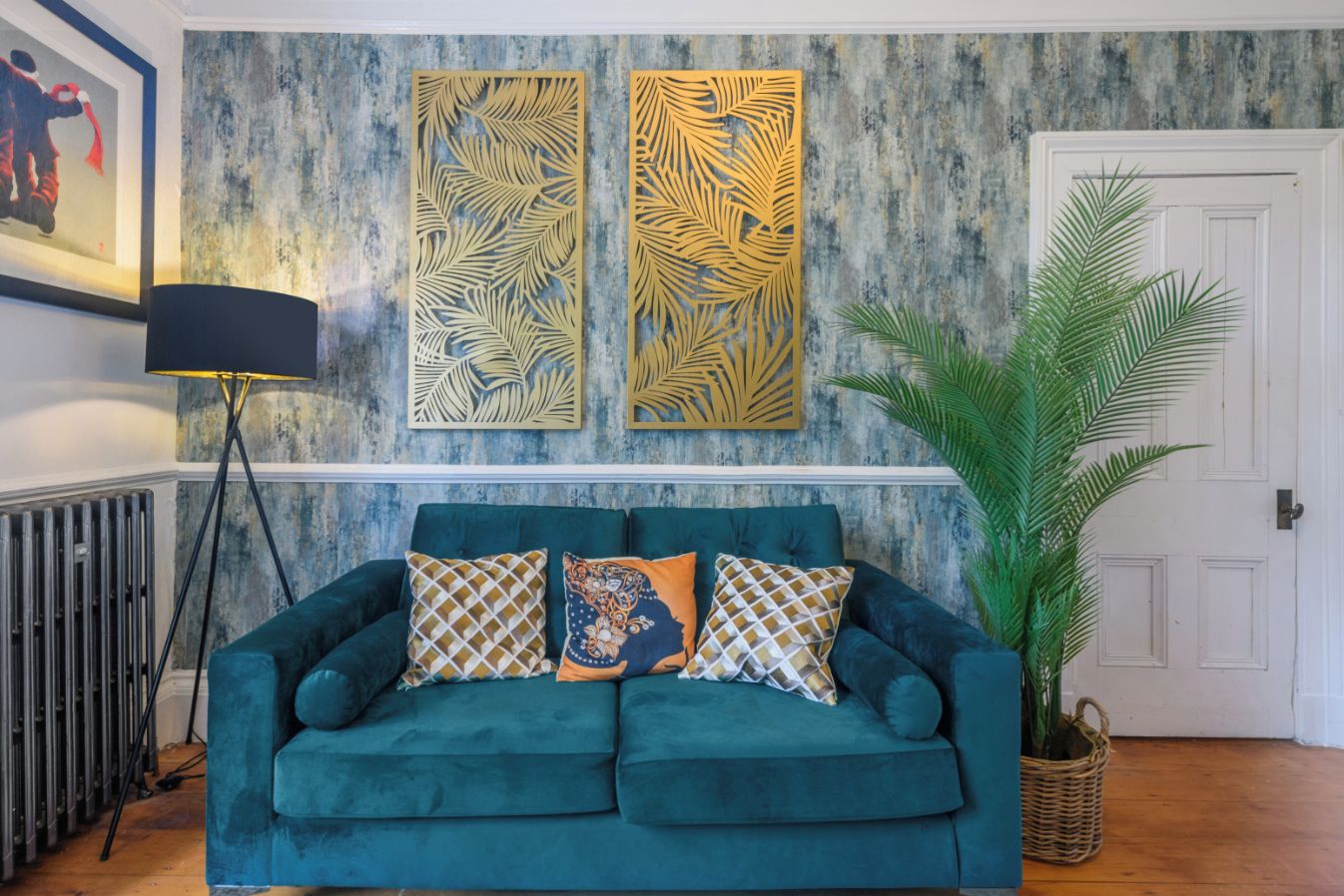 Un canapé de couleur turquoise agrémenté de coussins à motifs, un lampadaire et une grande plante en pot dans un salon doté d’un mur rustique et d’une porte blanche.