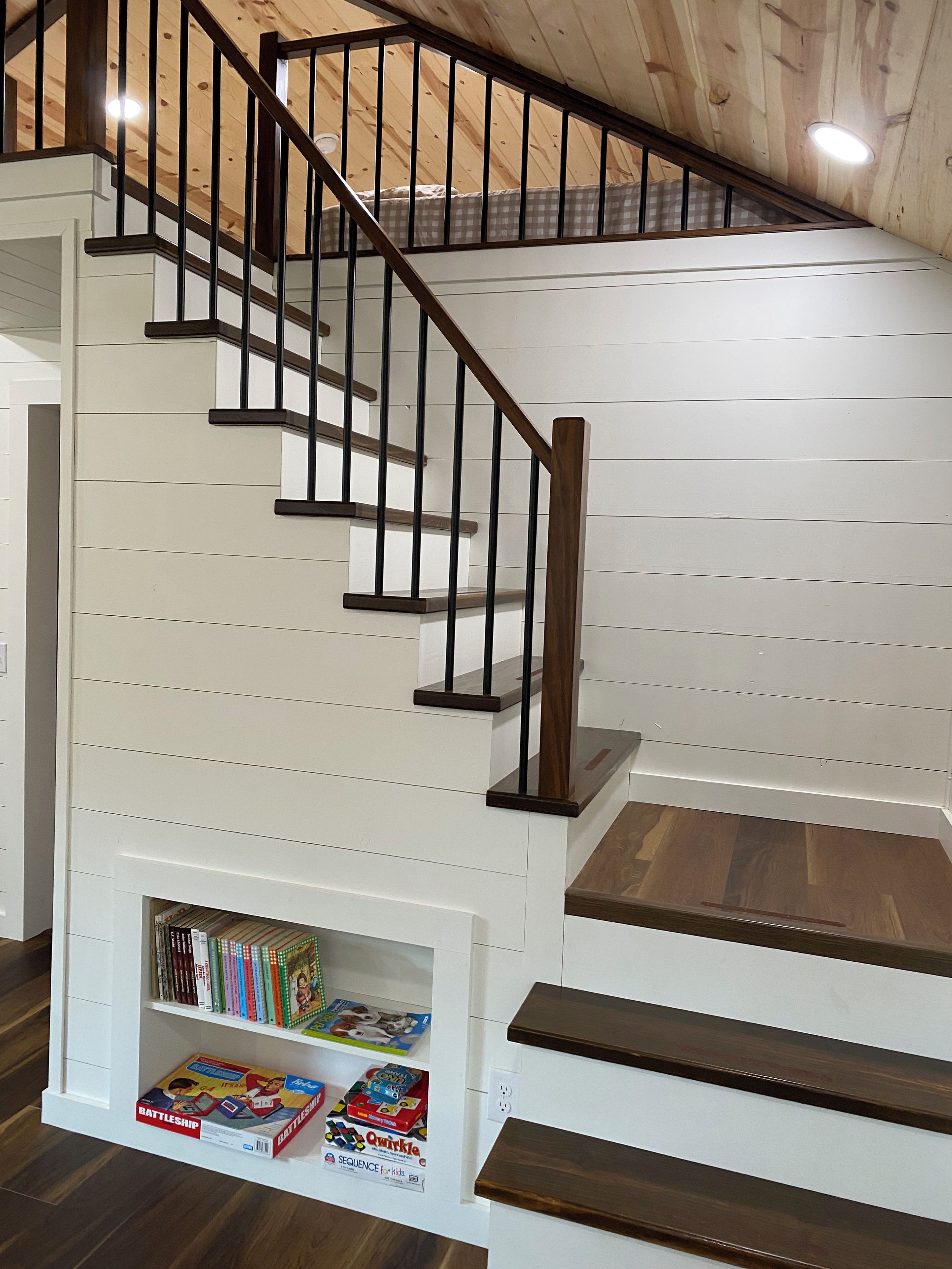 Un escalier avec des marches en bois et des rampes noires, menant à une mezzanine dotée d’un plafond en pente.