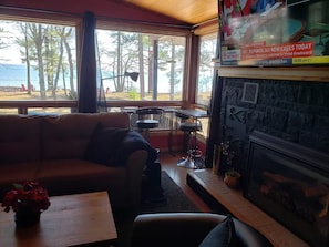 Une pièce chaleureuse avec une grande fenêtre donnant sur une plage, une table en bois, un canapé et une cheminée agrémentée d’un panneau décoratif.