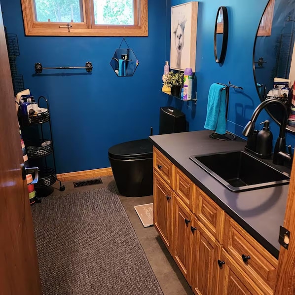 Une salle de bain avec un lavabo noir, des meubles en bois, un porte-serviettes, une poubelle noire et une fenêtre avec un cadre en bois.