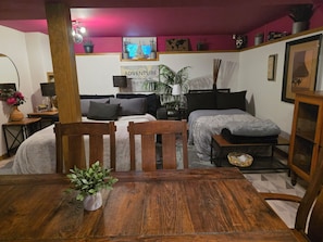 Une pièce confortable avec un lit, une table en bois et une chaise. On y trouve des plantes, une lampe et une étagère avec des objets décoratifs.