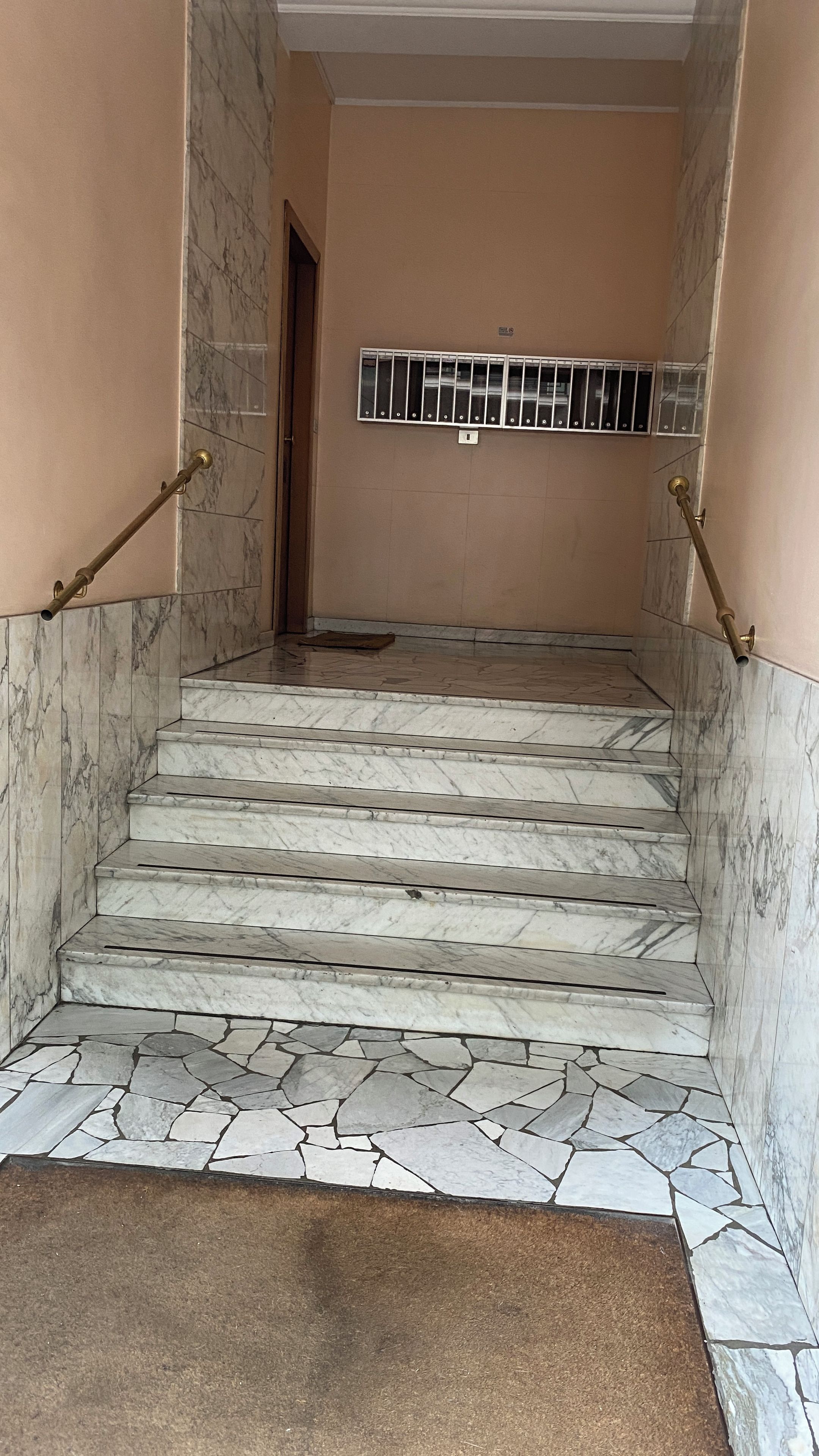 A marble staircase with a stone-tiled landing area, leading to a door with a metal grille.