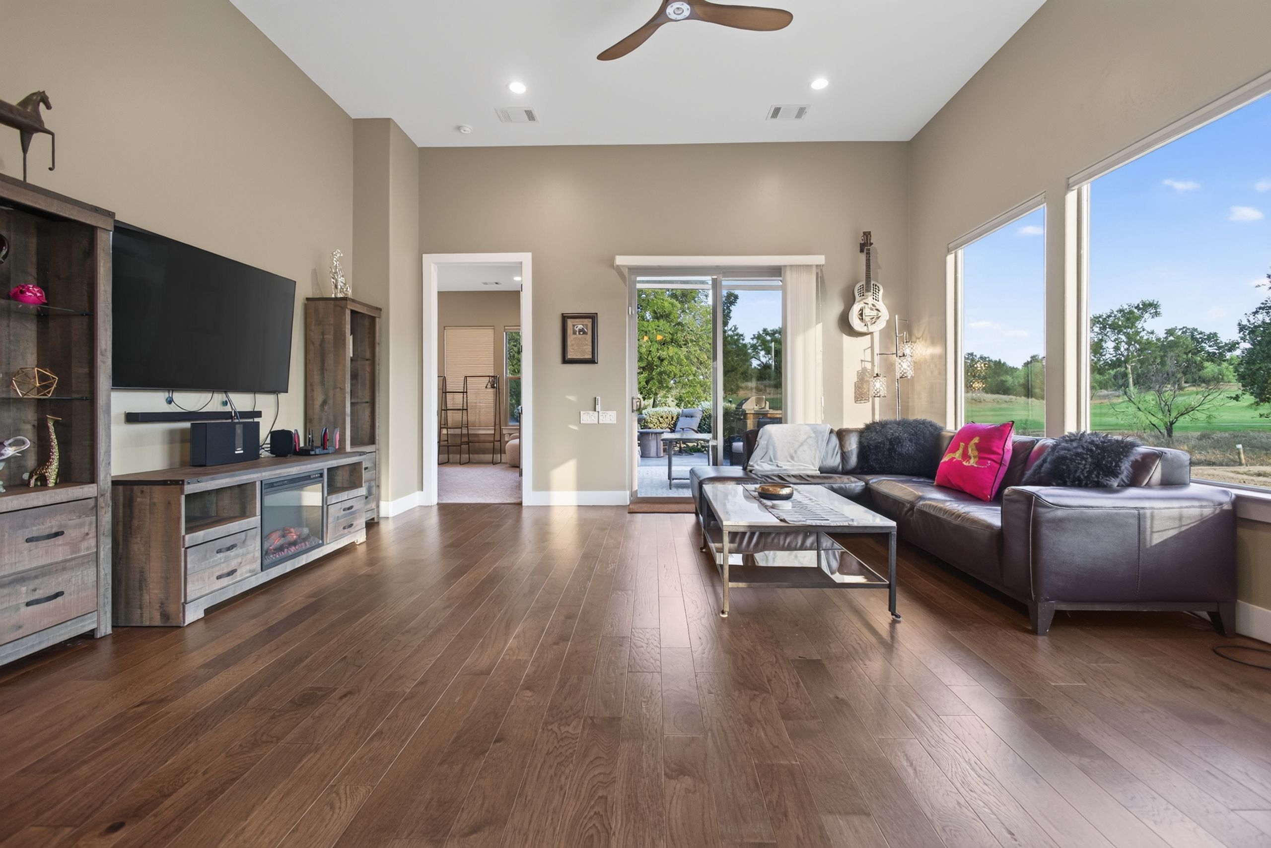 A spacious living room with a flat-screen TV, a sofa, and a coffee table. There is a large window providing a view of the outdoors.