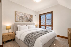 Ein Schlafzimmer mit einem großen Bett, einem Nachttisch, einer Lampe, einem Fenster mit Blick auf Bäume und einem Bild an der Wand.