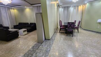 Una habitación de hotel con zona de estar, una mesa de comedor con sillas, una mesita blanca pequeña y una mesita auxiliar con superficie de vidrio.
