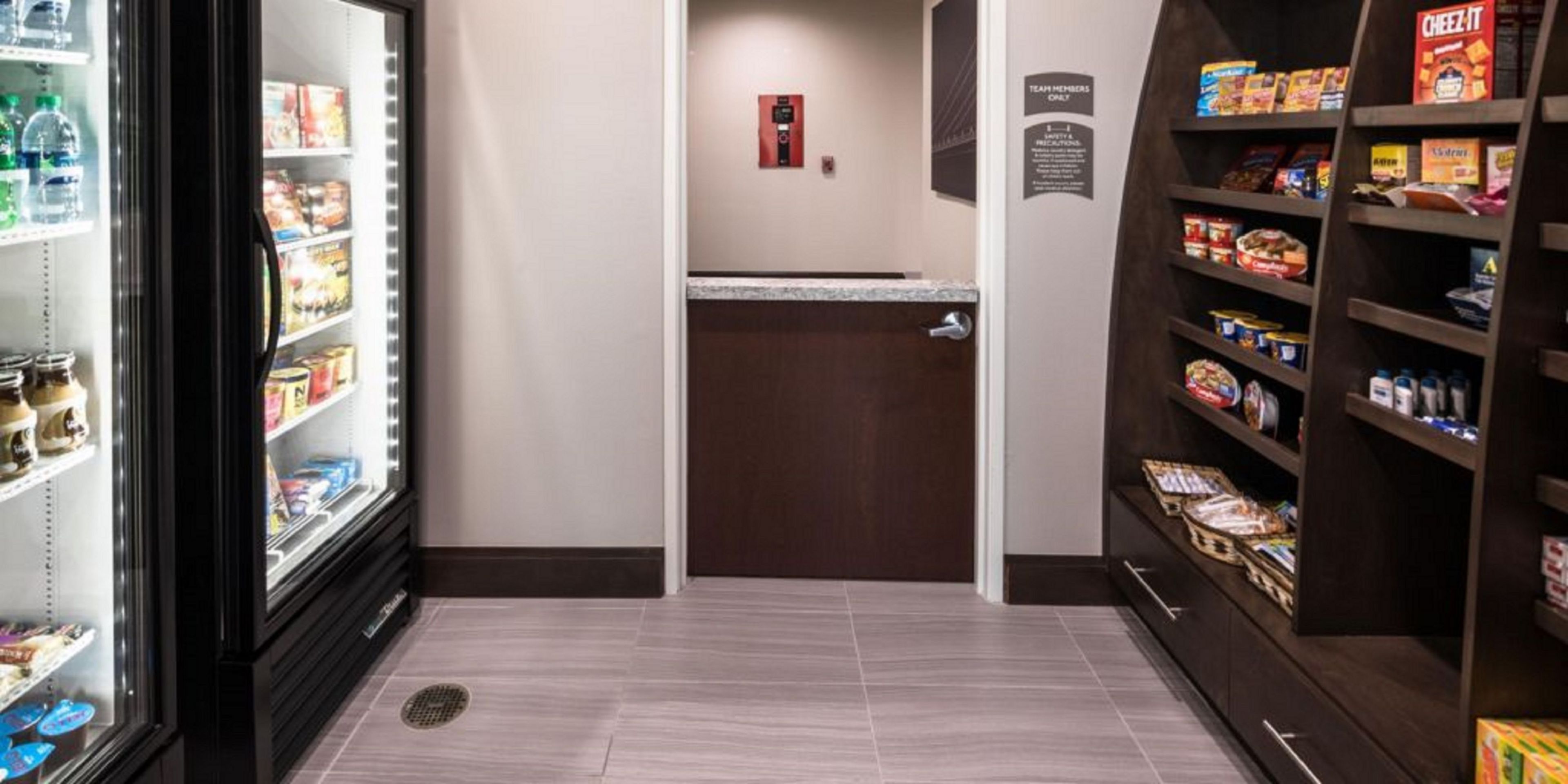 A hotel pantry with shelves stocked with snacks and beverages, a refrigerator, and a door leading to another room.