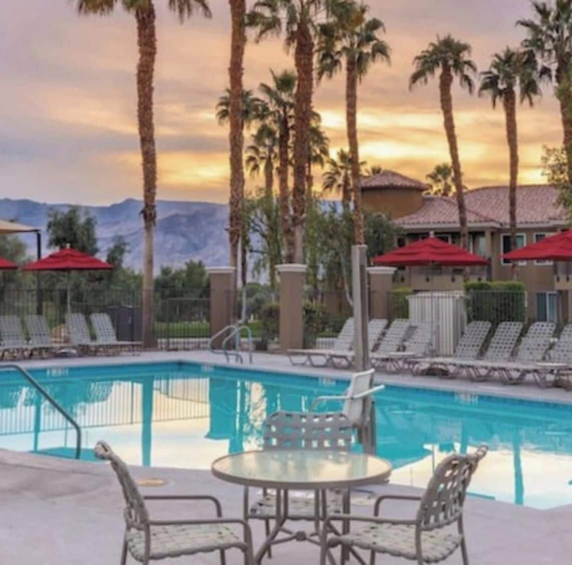 A pool area with lounge chairs and umbrellas, surrounded by palm trees and a mountain backdrop.