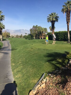 Ein Golfplatz mit blauem Himmel, Palmen und einem Bergpanorama im Hintergrund.