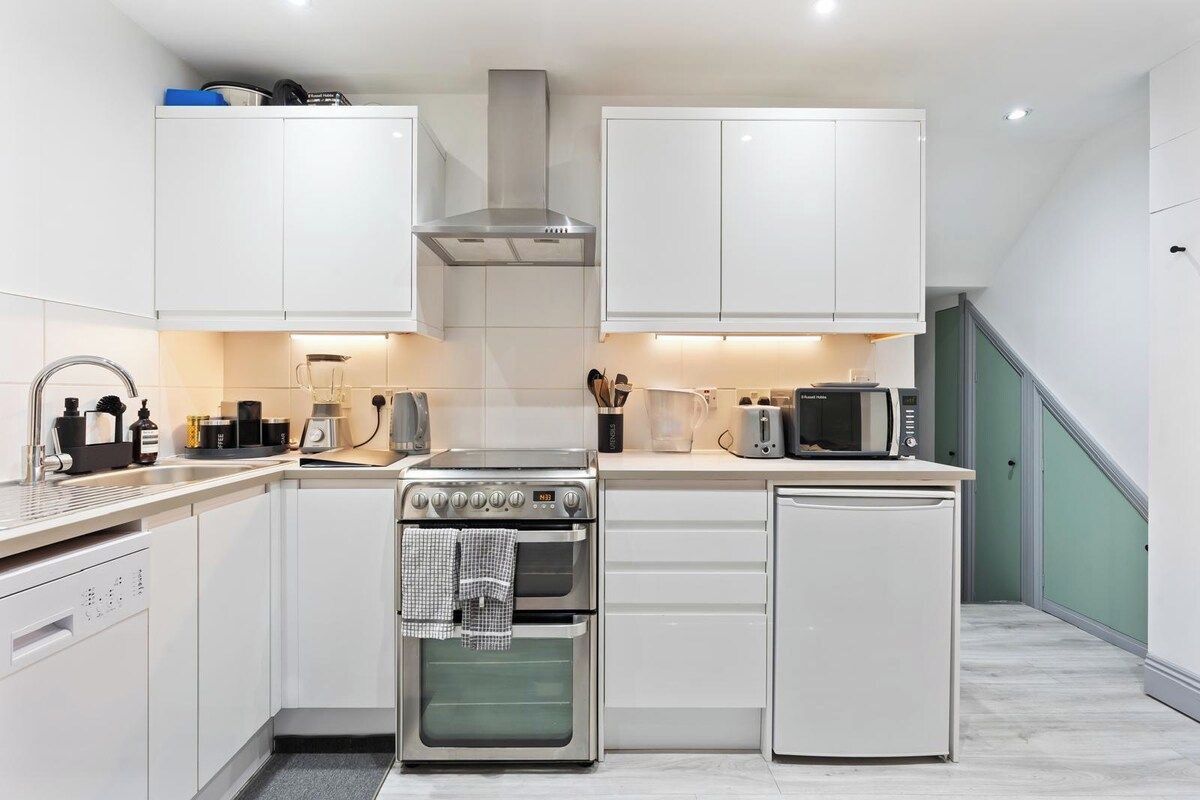 Cocina compacta con muebles blancos, extractor de acero inoxidable, horno empotrado, microondas y un refrigerador pequeño.