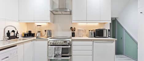Cocina compacta con muebles blancos, extractor de acero inoxidable, horno empotrado, microondas y un refrigerador pequeño.