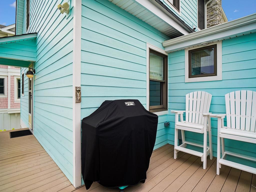 A covered grill on a wooden deck, two white Adirondack chairs, and a turquoise house with a window.