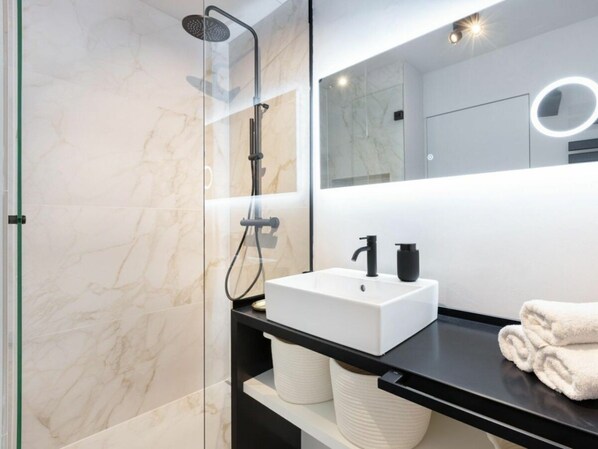 A modern bathroom with a glass-enclosed shower, a white sink with black fixtures, and a mirror with a circular light. - Sunset Loft Holiday Apartment (Sylt)