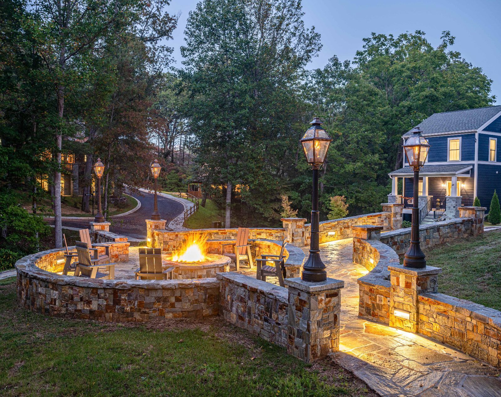 A stone fire pit area with benches, surrounded by a stone wall, and a house with a porch in the background.