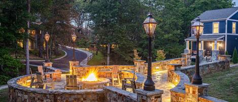 A stone fire pit area with benches, surrounded by a stone wall, and a house with a porch in the background.