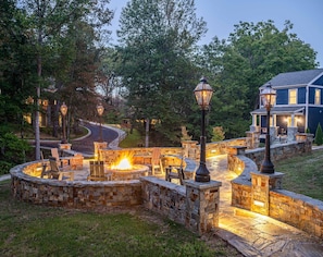 A stone fire pit area with benches, surrounded by a stone wall, and a house with a porch in the background.