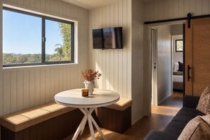 Ein Zimmer mit einem runden Tisch, einer Bank, einem an der Wand befestigten Fernseher und Blick durch ein Fenster.