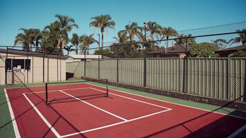 An outdoor tennis court with a net, surrounded by a chain-link fence, palm trees, and buildings in the background.