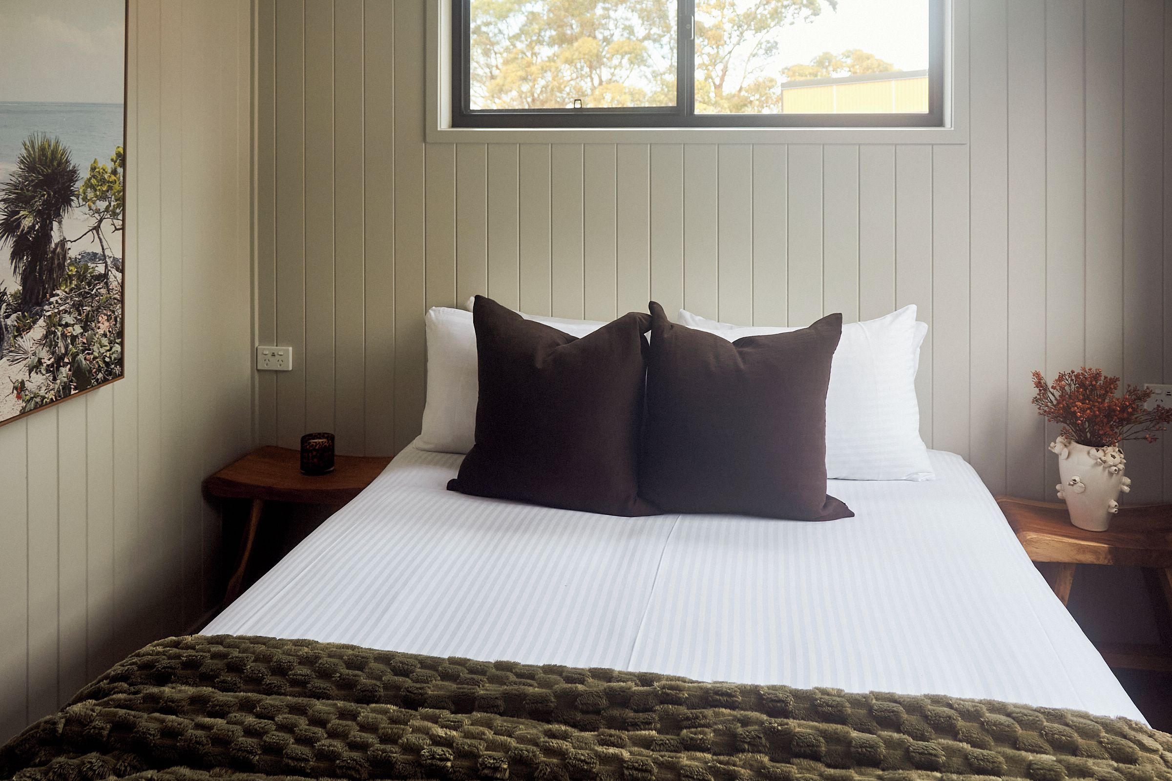 A bedroom with a large bed, a window with a view of trees, a nightstand with a cup, and a wall-mounted power outlet.