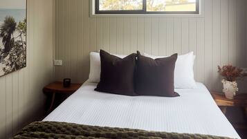 A bedroom with a large bed, a window with a view of trees, a nightstand with a cup, and a wall-mounted power outlet.
