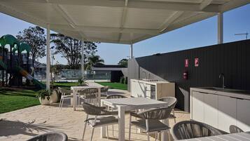 An outdoor patio area with tables and chairs, a barbecue station, and a children's play structure in the background.