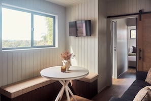 Una habitación con una mesa redonda, asientos tipo banco, un televisor de pantalla plana montado en la pared y vistas a través de una puerta abierta.