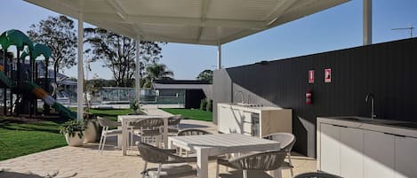 An outdoor patio area with tables and chairs, a barbecue station, and a children's play structure in the background.