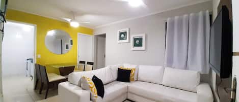 A modern living room with a white sectional sofa, a dining table, chairs, a ceiling fan, and a flat-screen TV.