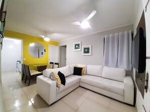 Uma sala de estar moderna com um sofá branco modular, uma mesa de jantar, cadeiras, um ventilador de teto e uma televisão de tela plana.