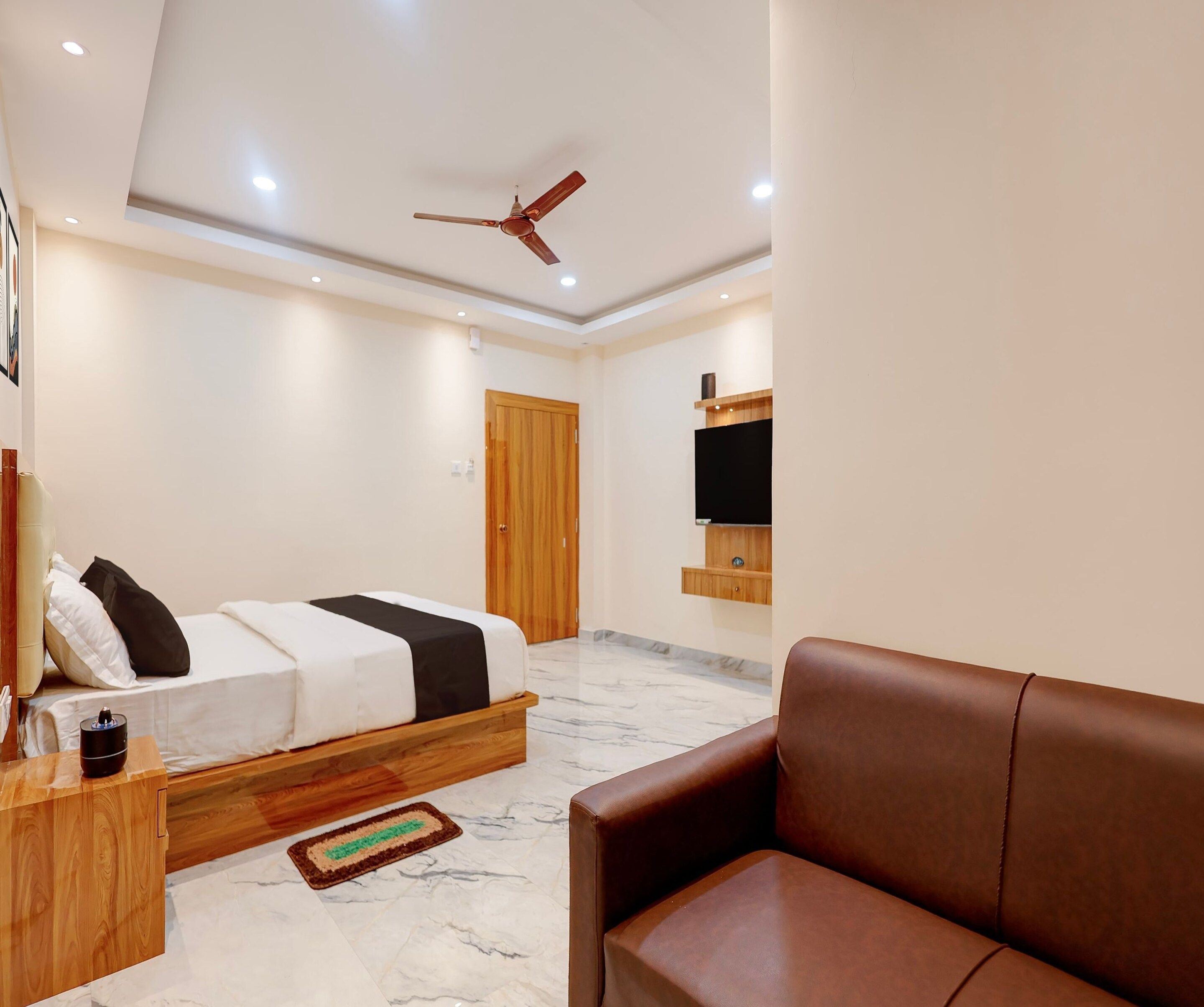 Ein modernes Schlafzimmer mit Bett, einem hölzernen Nachttisch, einem braunen Ledersofa, einem Deckenventilator und einem an der Wand montierten Fernseher.