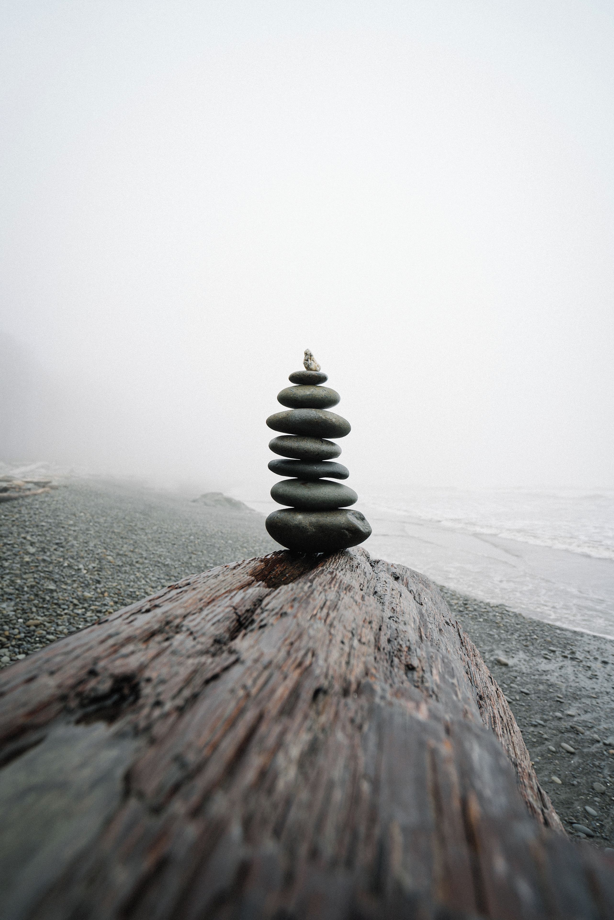 Ein Stapel glatter Steine, ausbalanciert auf einer verwitterten Holzoberfläche, mit einem unscharfen Strand und Meer im Hintergrund.
