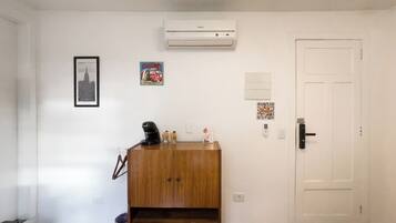 A room with a white door, a wall-mounted air conditioner, a framed picture, a small cabinet, and a trash can.