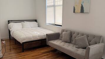 A bedroom with a bed, a sofa, a window with blinds, and a painting on the wall.
