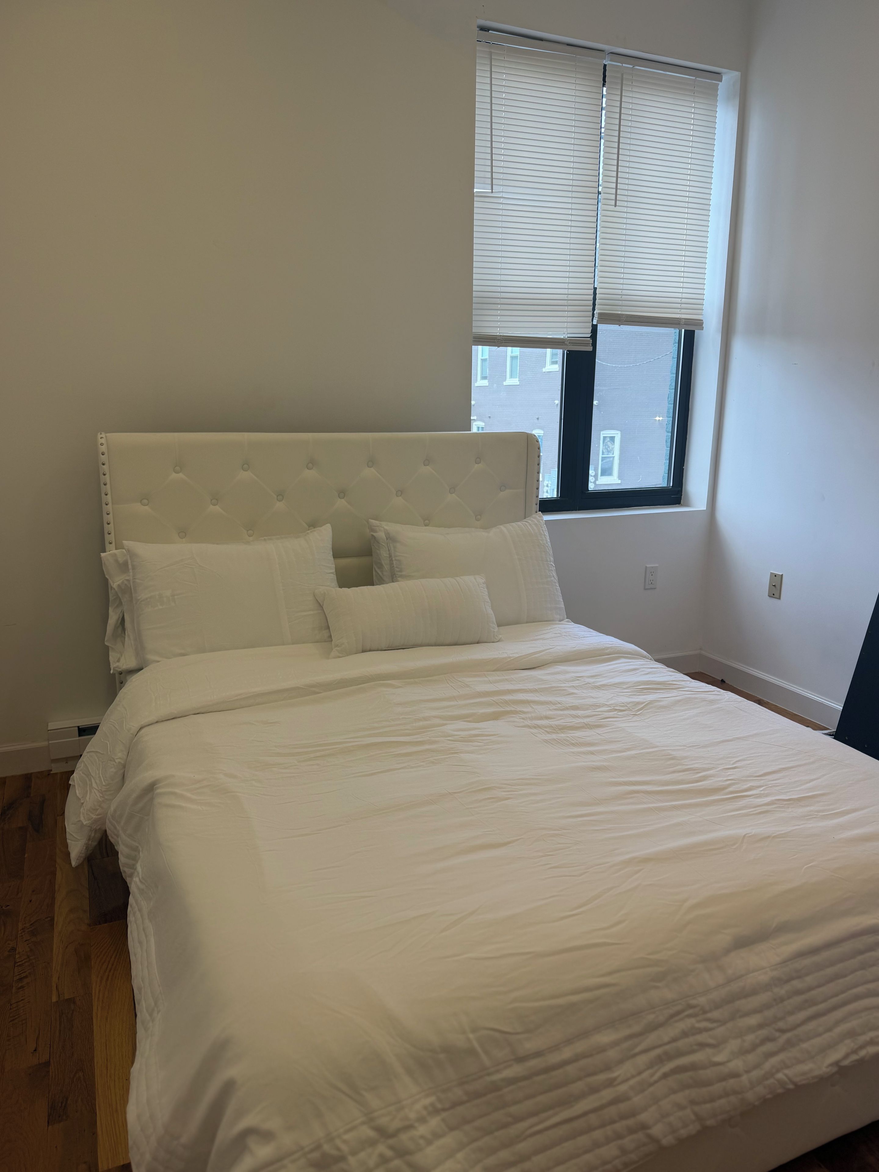 A neatly made bed with a tufted headboard and white bedding in a room with a window and plain walls.