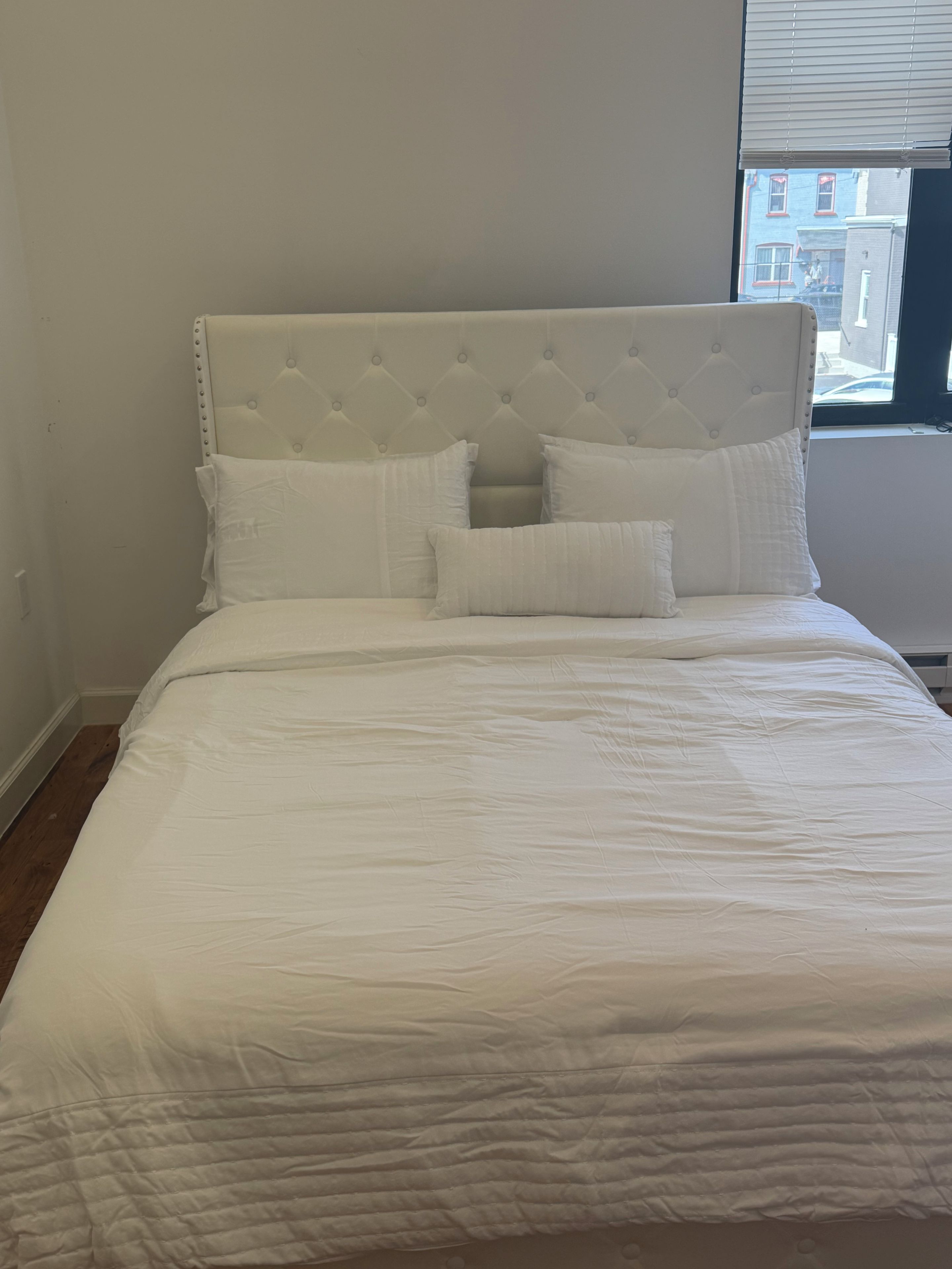 A bed with a tufted headboard, white bedding, and a window with blinds.