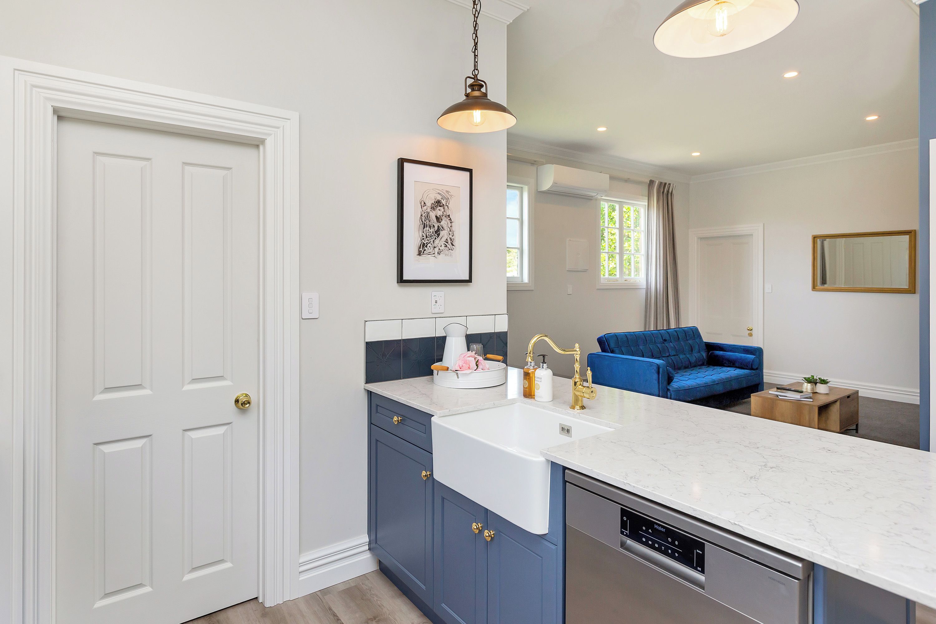 A modern kitchen with a white countertop, blue cabinets, and a dishwasher. There is a blue sofa in the adjacent living area.