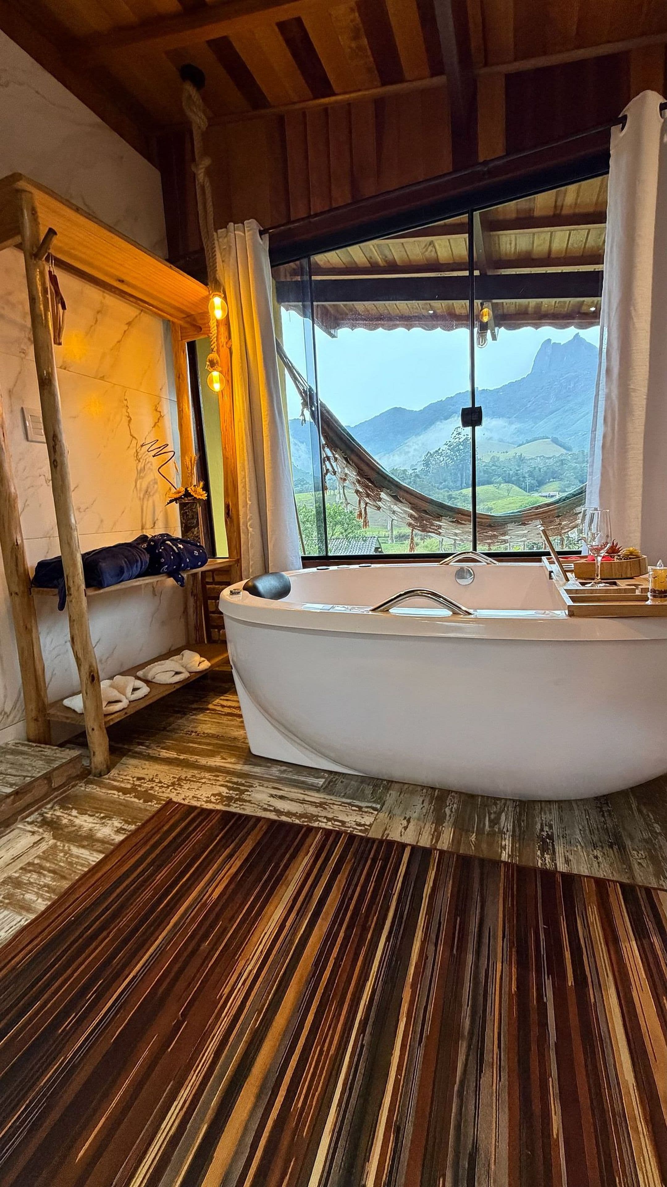 A room with a large bathtub, wooden flooring, a window with a view of mountains, and a hanging chair.