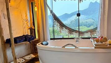 A room with a large bathtub, wooden flooring, a window with a view of mountains, and a hanging chair.