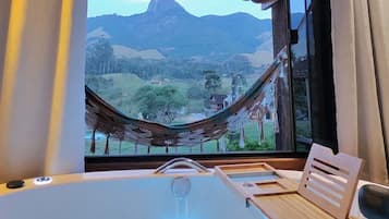A room with a large bathtub, a wooden chair, and a view of a mountain through a window with a hammock.