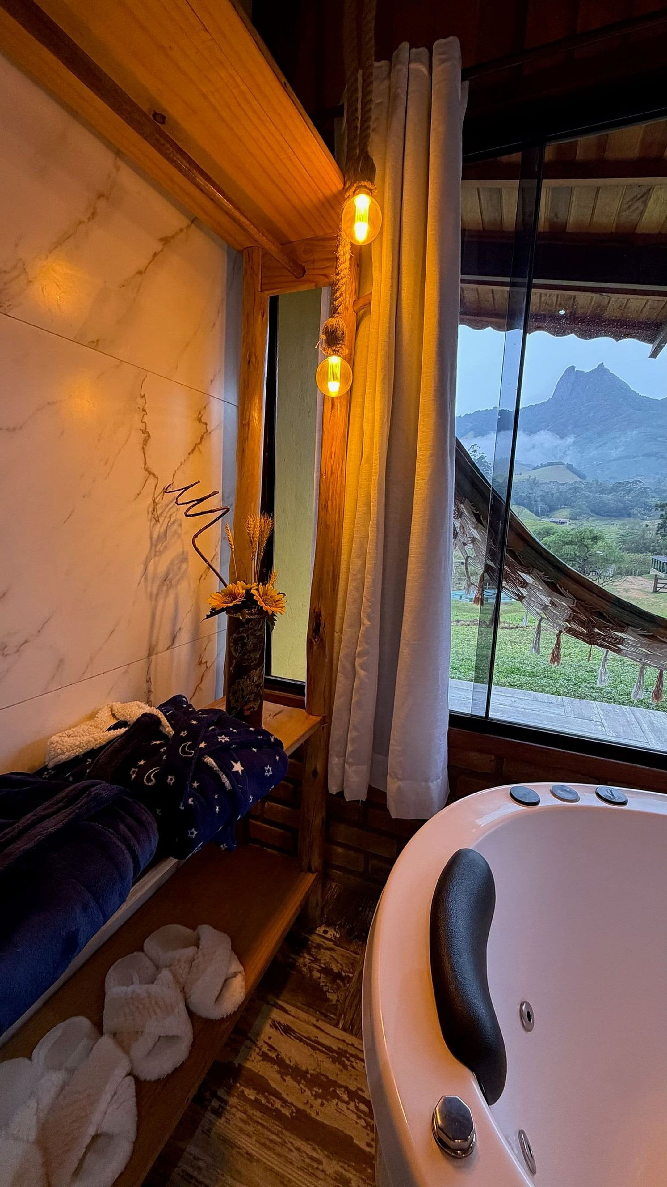 Una habitación con techo de madera, un ventanal con cortinas, cama con manta de diseño estrellado, bañera y un florero con flores.