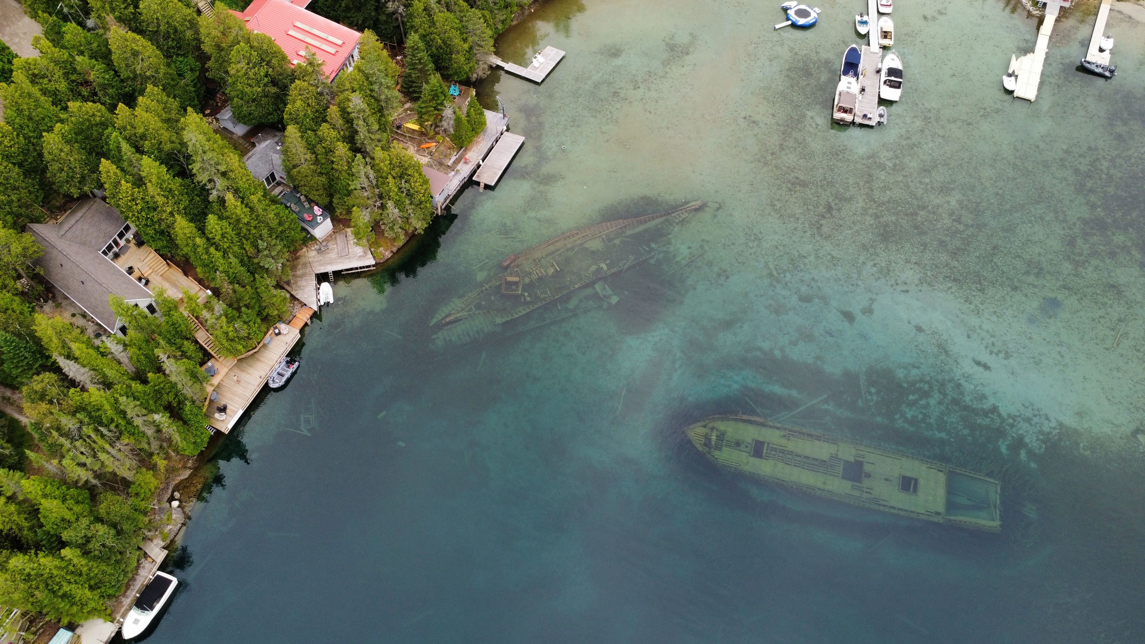 Vue aérienne d’une zone côtière avec un quai, des bateaux et un épave immergée dans une eau claire.