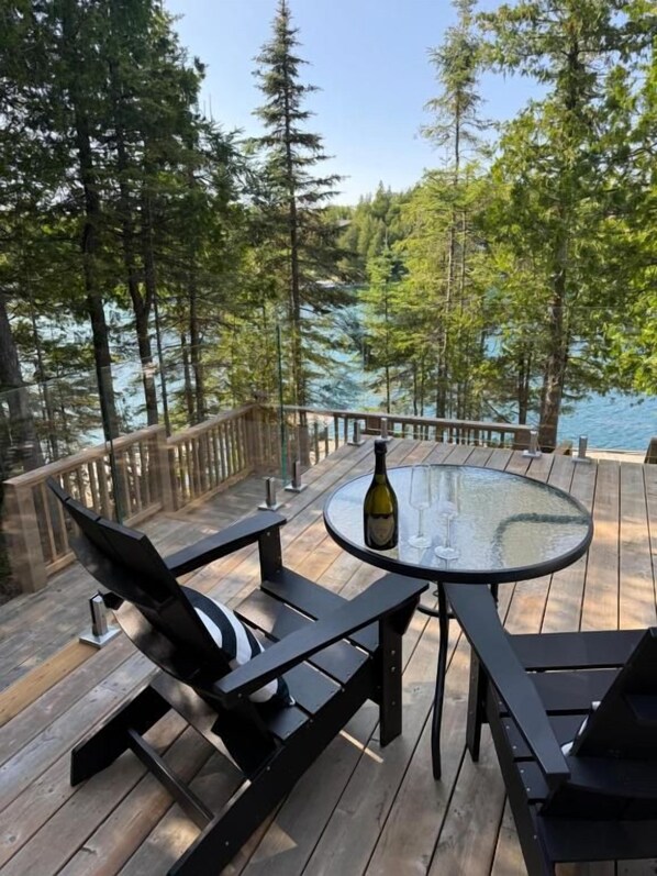 A wooden deck with a round table and chairs, overlooking a body of water and surrounded by trees.