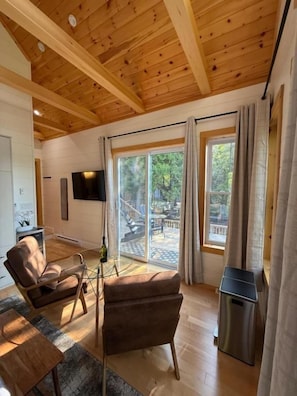 A room with wooden ceiling beams, a sofa, a small table, a TV mounted on the wall, and large windows with curtains.