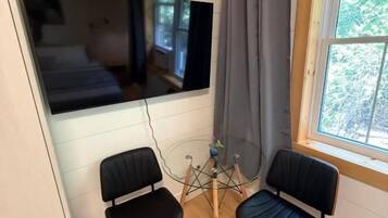 Une pièce dotée d’une grande fenêtre, de deux chaises noires, d’un plancher en bois et d’un téléviseur fixé au mur.