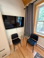 Una habitación con un ventanal grande, dos sillas negras, piso de madera y un televisor montado.