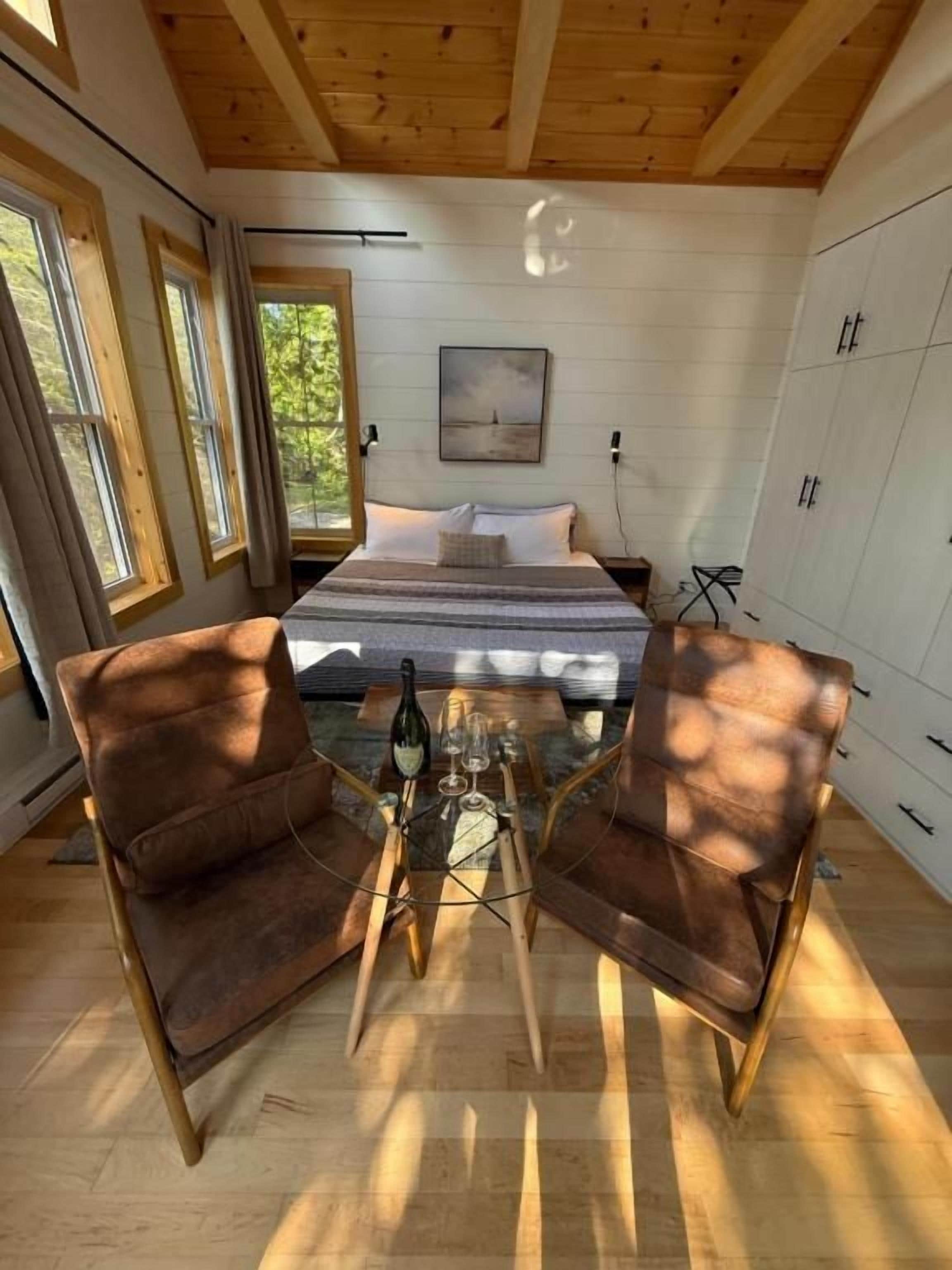 A bedroom with a bed, two armchairs, a glass-top coffee table, and a view of the outdoors.