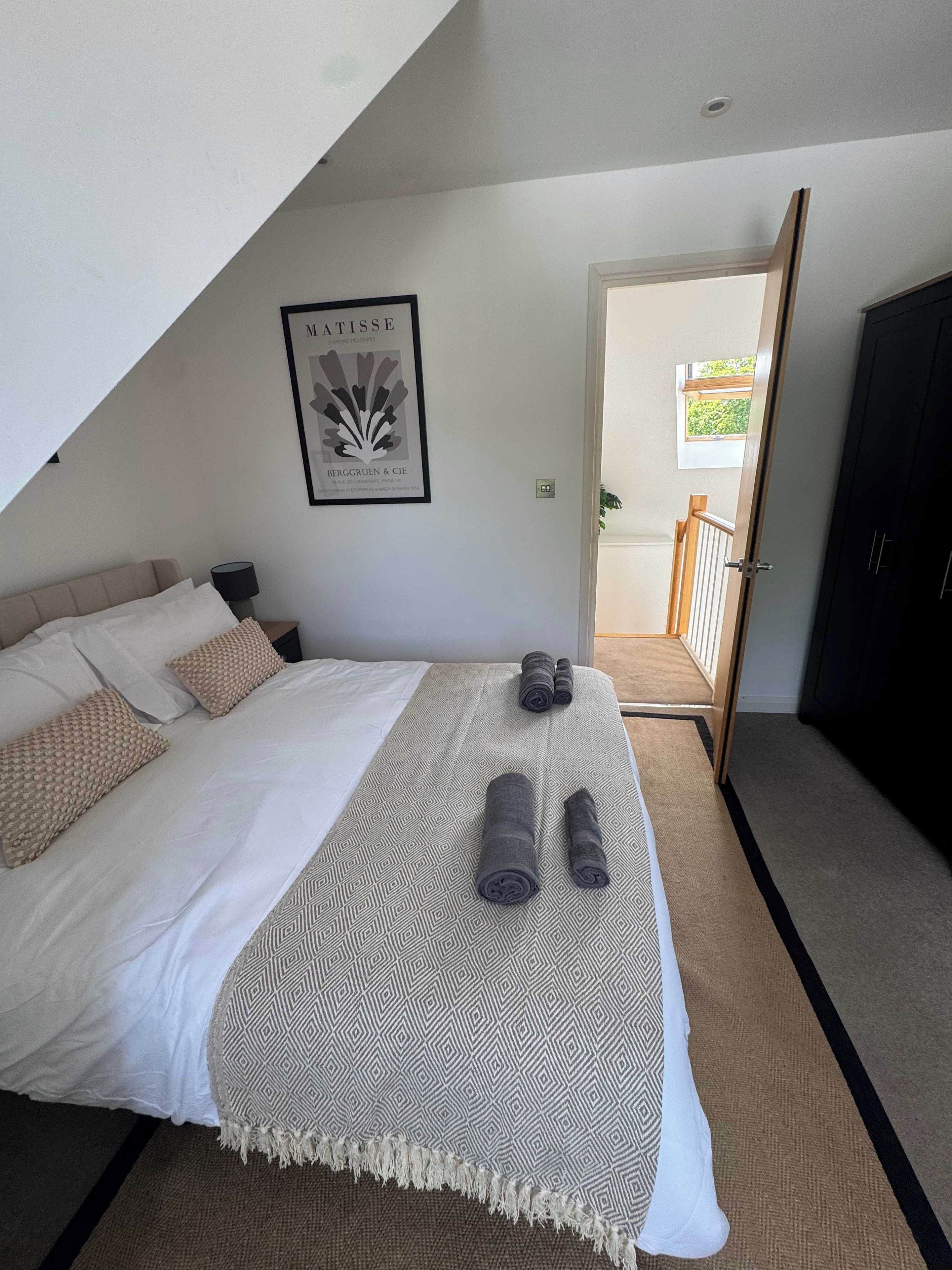 Una camera da letto con un letto, cuscini, una coperta e asciugamani arrotolati. C'è un quadro appeso al muro e una porta che dà su un balcone.