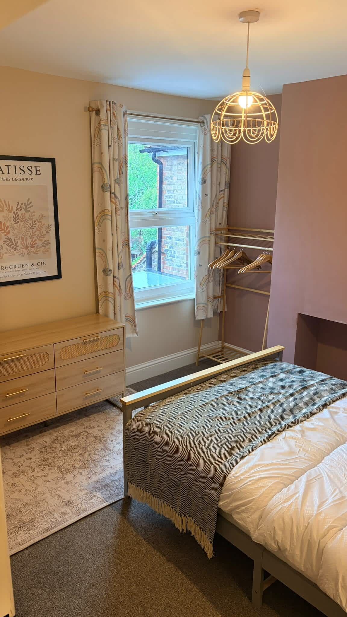 A bedroom with a bed, a dresser, a window with curtains, a hanging light fixture, and a framed picture on the wall.